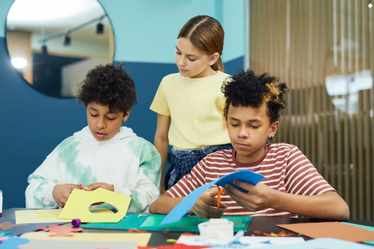 Two kids are cutting colored paper with scissors while a third child looks over their shoulders.